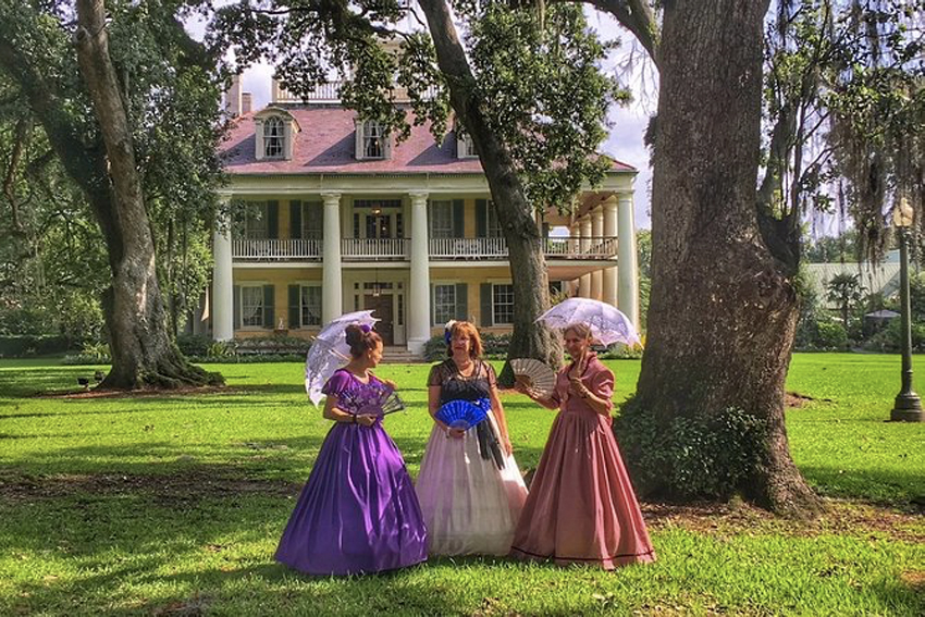 three women with sun umbrellas dressed in old fashioned long dresses in front of the southern mansion